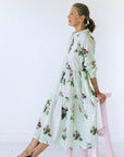 Floral print pale green midi dress, worn with black brogues. Model sitting on a pink stool and facing the left