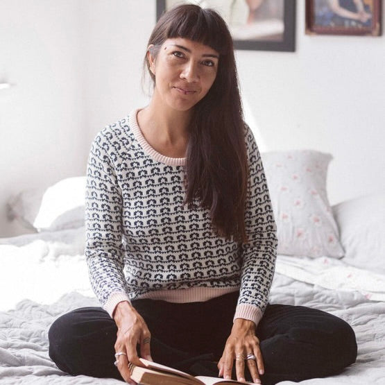 A woman sits on a bed with a book wearing the Tilda Jumper in Loop - Organic Cotton. The jumper has a pale pink neckline, cuffs and hem with the main pattern being black and white loops.