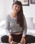 A woman sits on a bed with a book wearing the Tilda Jumper in Loop - Organic Cotton. The jumper has a pale pink neckline, cuffs and hem with the main pattern being black and white loops.